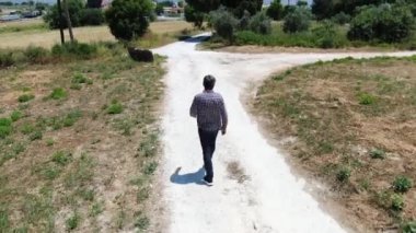 Kot pantolon, ekose gömlek ve güneş gözlüğü giyen bir adam yukarıdan Video bir ülke yol boyunca yürürken. Dron bir adamı takip ediyor. İhA'dan ateş etme.