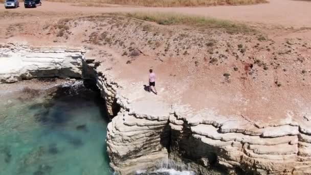 Férfi öltözött nadrág, cipők és a póló egy szikla mentén sétált. Habos hullámok üti a sziklás parton. Sportlövészet a drone. Ciprus. — Stock videók