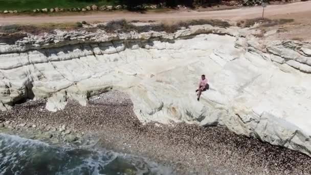Man gekleed in shorts, sneakers en een T-shirt zittend op een klif. Schuimige golven die de rotsachtige kust raken. Schieten vanaf de drone. Cyprus. — Stockvideo