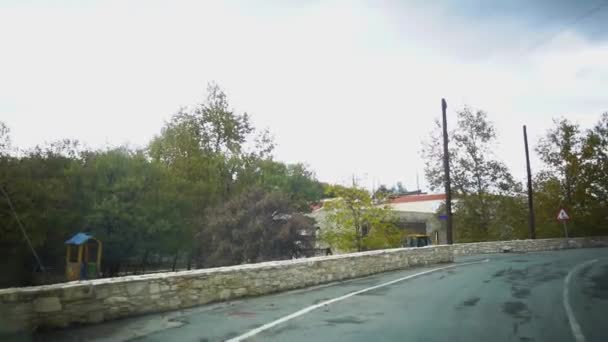 Vista desde un coche en movimiento en una calle estrecha con casas, truchas y señales de tráfico en un día nublado después de la lluvia. Chipre . — Vídeo de stock