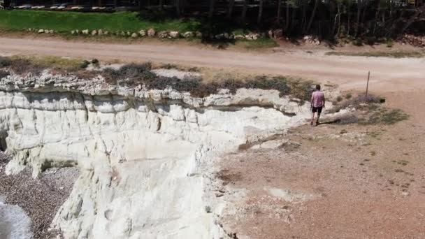 Férfi öltözött nadrág, cipők és a póló egy szikla mentén sétált. Habos hullámok üti a sziklás parton lement a vízbe. Sportlövészet a drone. Ciprus. — Stock videók