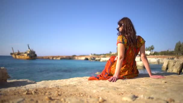 Hermosa joven con un vestido sentado junto a la costa rocosa en el fondo de la pintoresca vista de un viejo barco, cielo y acantilados. Chipre . — Vídeo de stock