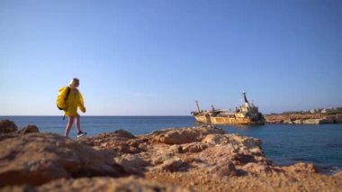 Güzel genç kadın turist sarı yağmurluk pitoresk deniz, eski gemi ve mavi gökyüzü arka planda kayalık plaj tarafından yürüyüş. Kıbrıs. Yavaş çekim.