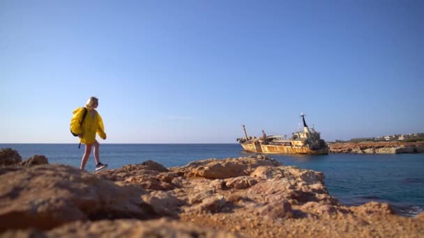 Hermosa joven turista en impermeable amarillo caminando por la playa rocosa en el fondo del mar pintoresco, barco viejo y cielo azul. Chipre. Movimiento lento . — Vídeos de Stock