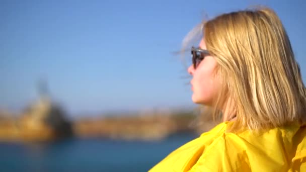 Primer plano de una chica con el pelo rubio usando gafas de sol y un impermeable amarillo de pie en un acantilado en tiempo ventoso y soleado. En cámara lenta. Chipre . — Vídeo de stock