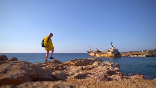 Hermosa joven turista en impermeable amarillo caminando por la playa rocosa en el fondo del mar pintoresco, barco viejo y cielo azul. Chipre. Movimiento lento . — Vídeos de Stock