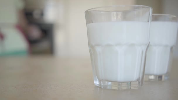 Twee glazen melk op de tafel op de achtergrond een jonge vrouw die vers gebakken broodjes uit de oven trekt. Close-up. Advertising concept. — Stockvideo