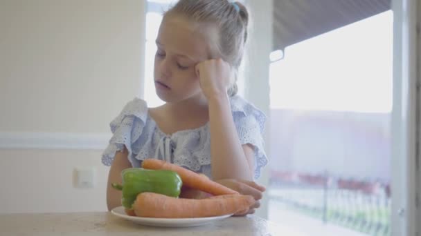 Nešťastně naštvaná holčička před talířkou se zeleninou v kuchyni. Dítě odtlačí talíř s nechutná potravou. Ten kluk odmítá jíst pepř a mrkev — Stock video