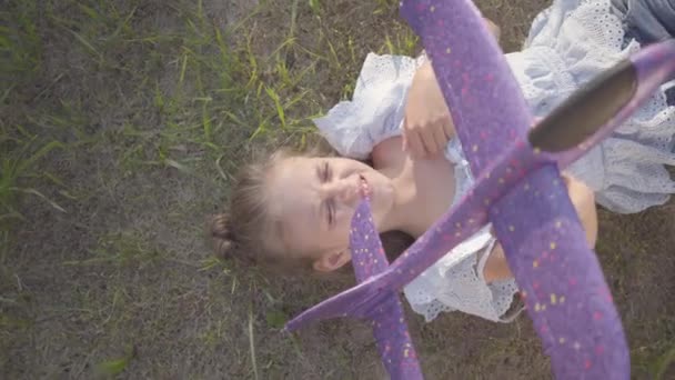 Kleines Mädchen an einem Sommerabend in den Ferien unter der Sonne im Gras liegend und ein Spielzeugflugzeug in der Hand haltend. — Stockvideo