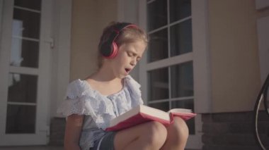 Yaz tatili sırasında sevimli kız müzik dinleme, şarkı ve kitap okuma evin yakınındaki avluda oturan