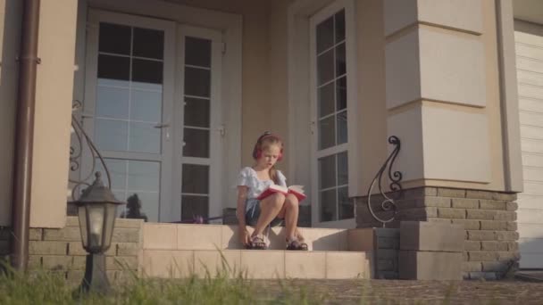Aranyos lány a nyári szünetben ül az udvaron ház közelében hallgat zenét, éneklés és olvasás könyv — Stock videók