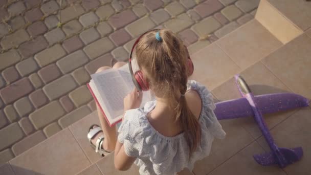 Bastante linda niña en auriculares leyendo el libro, el pequeño avión acostado en el porche. El niño pasa tiempo al aire libre en el patio trasero. Una infancia sin preocupaciones. Vista trasera . — Vídeo de stock