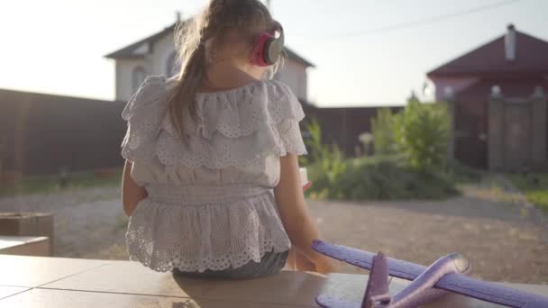 Kitap okuyan kulaklıklı sevimli küçük bir kızın arka görünümü, verandada yatan küçük bir uçak. Çocuk arka bahçede dışarıda vakit geçiriyor. Kaygısız çocukluk — Stok video