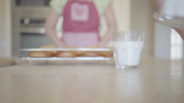 Mano bruscamente mette un bicchiere con il latte sul tavolo, versandolo sul tavolo primo piano. Donna irriconoscibile in grembiule con torte appena sfornate sullo sfondo. Pranzo in una famiglia felice — Video Stock