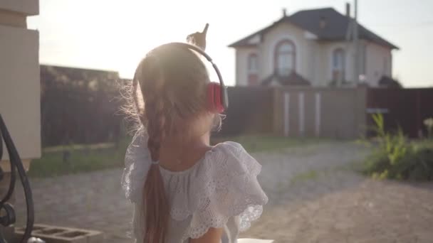 Вид сзади на симпатичную маленькую девочку в наушниках, читающую книгу, сидящую на крыльце и машущую руками. Ребенок проводит время на улице на заднем дворе. Беззаботное детство — стоковое видео