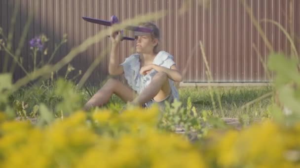 フェンスの下の草の上に座って小さな飛行機で遊んでかわいいかわいい女の子の肖像画。子供は裏庭で屋外で時間を過ごす。気楽な子供時代 — ストック動画