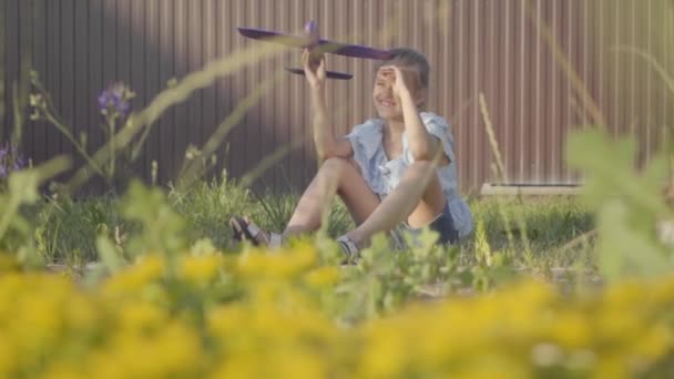 Entzückendes Mädchen mit einem Spielzeugflugzeug, das auf dem grünen Gras im Hof sitzt. Mädchen haben Spaß im Freien. Unbeschwerte Kindheit. Zeitlupe — Stockvideo