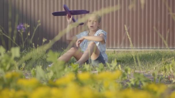 Portrait of a pretty cute little girl launching the small plane sitting on the grass under the fence. The child spending time outdoors in the backyard. Carefree childhood. Slow motion — Stock Video