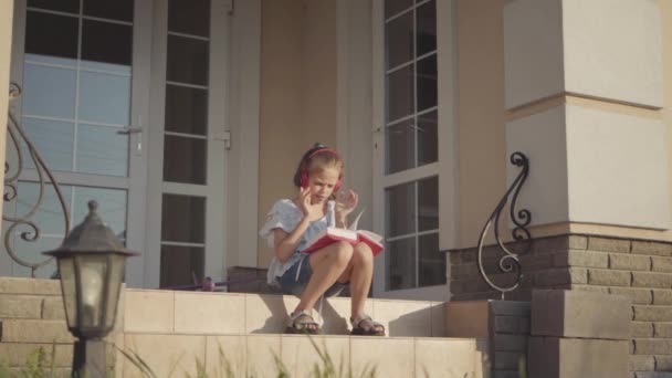 Adorable chica durante las vacaciones de verano sentado en el patio cerca de la casa escuchando música, cantando y leyendo libro. Movimiento lento . — Vídeos de Stock