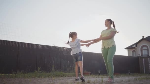 Happy little cute girl and her mother spinning around holding hands in the backyard. The woman and child having fun in front of shining sun. Mum and child spending time together outdoors. Slow motion — Stock Video