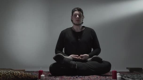 Красивый молодой человек сидит на полу с закрытыми глазами, свечи стоят рядом. Духовность, магия, концепция йоги. Камера движется вправо . — стоковое видео