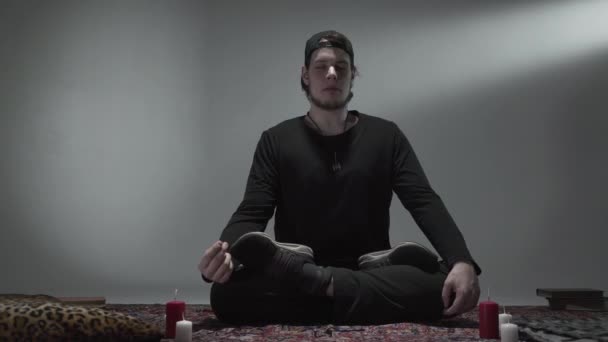 En ung hipster som sitter med lukkede øyne og har kryssben i et tomt rom med stearinlys som mediterer. Spiritualitet, magi, yogakonsept . – stockvideo