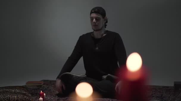 Jonge hipster zitten met gesloten ogen en kruis poten in een lege ruimte met kaarsen mediteren. Spiritualiteit, magie, yoga concept. — Stockvideo
