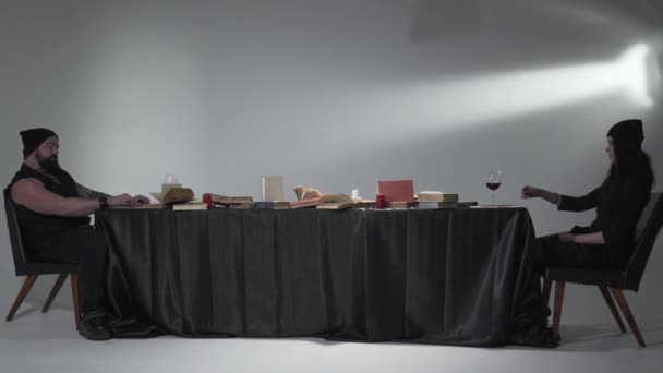 Scena insolita l'uomo barbuto brutale e la giovane donna in abiti neri seduti al lungo tavolo con molti libri su di esso che bevono vino rosso. Il concetto dello show. Studio girato — Video Stock