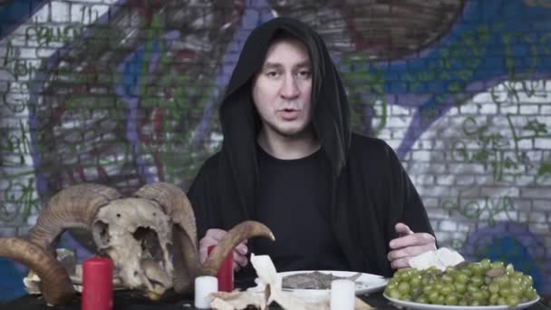 Podejrzany człowiek w czarnych ubraniach z kapturem na głowie siedzi przy małym stole przed świecami, winogrona i czaszka barana, rozmawiając patrząc w aparacie. Ciemny Wiedźmin obchodzi — Wideo stockowe