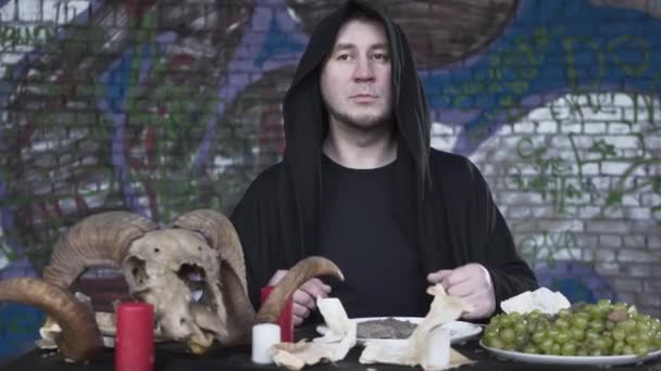 Mumlar, üzüm ve koç kafatası önünde küçük bir masada oturan baş kaputu ile siyah giysiler içinde Portre adam. Karanlık cadı kutlama. Stüdyo çekimi — Stok video