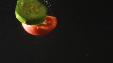 Salatalık ve domates dilimleri siyah arka plan üzerinde suya düşüyor. Kabarcıklar ile suda taze sebze. Organik gıda, sağlıklı yaşam tarzı. Yavaş çekim.