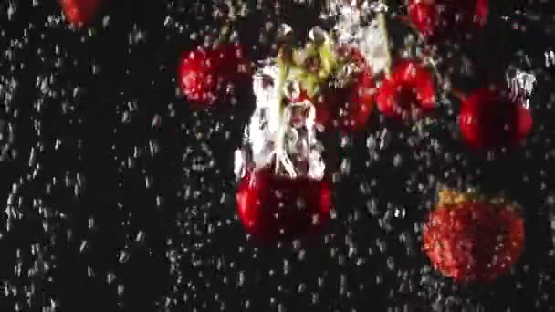 La caída de fresas frescas y cerezas salpicando en agua con gas sobre fondo negro. De cerca. — Vídeo de stock