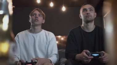 Oyun odasında kanepede oturan iki mutlu adam video oyunu oynuyor. Arkadaşlar içeride birlikte vakit geçiriyorlar. Video oyunu ve eğlence konsepti.