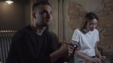 Portre genç adam bir dizüstü bilgisayarda yazarak sevimli smilling kız ile loft tarzı odada kanepede oturan video oyunu oynarken. Video oyunu ve eğlence konsepti.