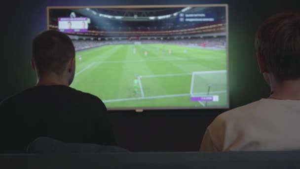 Två killar som spelar video spel i spelrummet sitter på soffan inomhus. Vänner som spelar fotboll. Vänner spenderar tid tillsammans. Video spel och fritid Concept. — Stockvideo