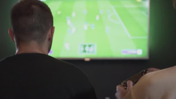 Widok z tyłu dwóch facetów gra w gry wideo w pokoju gier siedzi na kanapie. Znajomi grają w piłkę nożną lub piłkę nożną za pomocą joysticków. Koncepcja gry wideo i rozrywki. — Wideo stockowe