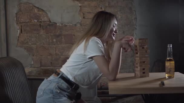 Mujer joven jugando a la torre de madera en la habitación de estilo loft. Ocio en interiores. Botella de cerveza de pie sobre la mesa. Chica despreocupada relajándose en la sala de juegos. Concepto de ocio en interiores — Vídeo de stock