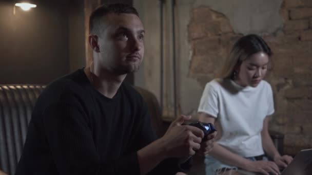 Retrato joven jugando el videojuego sentado en el sofá en la habitación de estilo loft con linda chica olorosa escribiendo en un ordenador portátil. Videojuego y concepto de ocio . — Vídeos de Stock