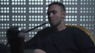 Portre kendine güvenen genç adam karanlık loft tarzı odada nargile sigara. Çekici adam kamera bakarak koltukta oturan duman nefes. İç mekanlarda boş zaman