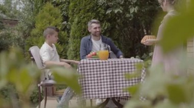 Baba, anne ve iki çocukları bir masada oturup bahçede güneşli bir günün tadını çıkarırken öğle yemeği yiyorlardı. Kadın ve kız pasta getiriyorlar. Yaz mevsimi, mutlu aile