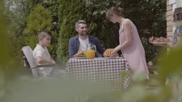 Papa, maman et leurs deux enfants assis à une table, déjeunant dans le jardin en profitant d'une journée ensoleillée. La femme et la fille apportent à manger. L'été, famille heureuse — Video