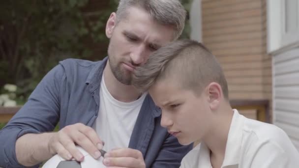 父亲和他的儿子坐在门廊上，手里拿着一个放气的足球特写。爸爸帮助男孩把球修好。家人在一起。夏季休闲 — 图库视频影像