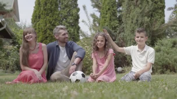 Porträt einer glücklichen Familie, die zusammen auf dem Gras im Garten sitzt. Mutter, Vater, Sohn und Tochter schauen in die Kamera. Freizeit im Sommer, unbeschwerte Kindheit — Stockvideo