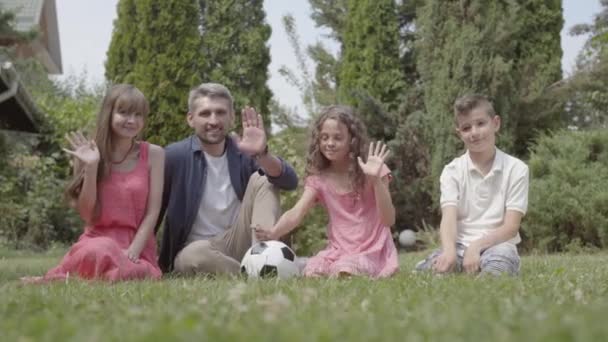 Portrait mignon famille heureuse assis sur l'herbe dans le jardin ensemble. Mère, père, fils et fille regardant dans la caméra en souriant et en agitant les mains. Loisirs d'été, enfance insouciante — Video