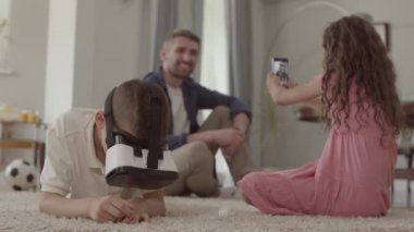 Kabarık halıda yerde yatan erkek ve kız, sanal gerçeklik gözlüğü kullanan kardeş, çocukların arka planında oturan babanın fotoğrafını çekiyor. Evde mutlu dost aile