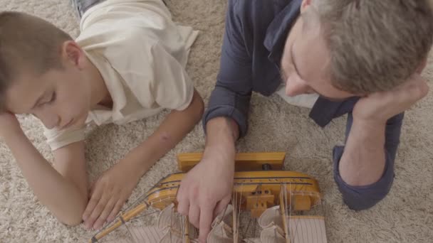 Portret schattige vader en zijn zoon liggen thuis op de vloer op het pluizige tapijt spelen met schip model close-up. Gezins tijd samen doorbrengen. Achteraanzicht — Stockvideo