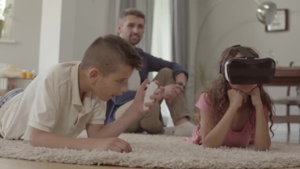 Мальчик и девочка лежат на полу на пушистом ковре в очках виртуальной реальности, отец сидит на заднем плане с детьми. Счастливая дружная семья дома — стоковое видео