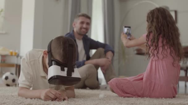 Мальчик и девочка лежат на полу на пушистом ковре, брат в очках виртуальной реальности, сестра фотографирует отца, сидящего на фоне детей. Счастливая дружная семья дома — стоковое видео