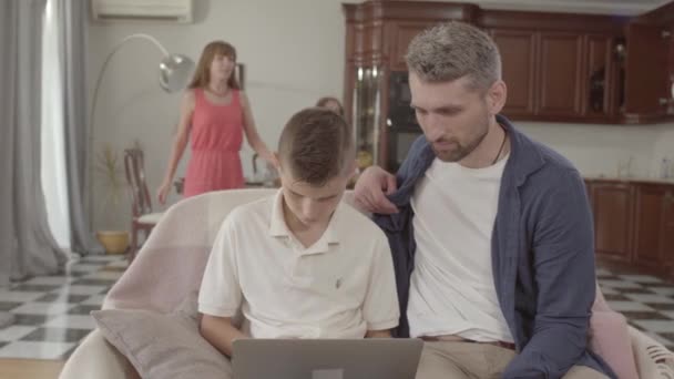 Fadern och sonen sitter tillsammans på fåtöljen med den bärbara datorn. Vacker mor och dotter kommer till dem och tittar på skärmen. Lycklig vänlig familj hemma — Stockvideo