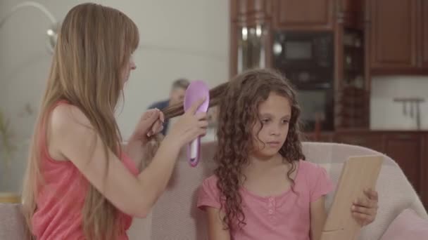 Genç anne ön planda kızının saçlarını fırçalarken, baba ve oğlu arka planda dizüstü bilgisayarda oynuyor. Mutlu aile evde birlikte vakit geçiriyor — Stok video